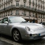 Porsche 911 argentée garée dans une rue parisienne, vue en angle avant trois quarts pour illustrer une assurance auto haut de gamme