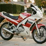 Yamaha TZR 50cc des années 90 garé dans une rue résidentielle, vue 3/4 avant mettant en valeur le carénage sportif et la livrée blanc et rouge